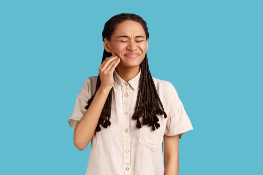 Displeased woman suffers from toothache, touches cheek, needs to see dentist, has health problems, closes eyes with pain, wearing white shirt. Indoor studio shot isolated on blue background.