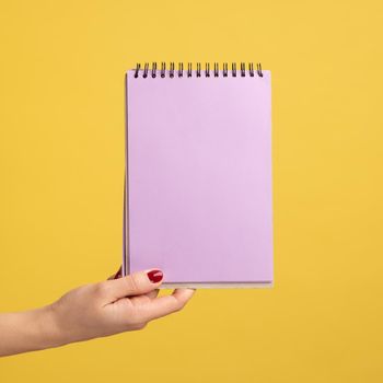 Profile side view closeup of woman hand holding purple notepad in hand and showing empty paper. Indoor studio shot isolated on yellow background.