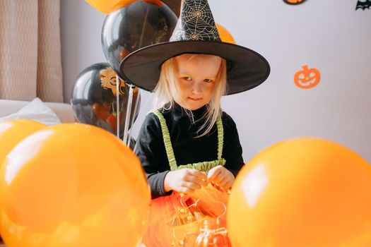 Children's Halloween - a girl in a witch hat and a carnival costume with airy orange and black balloons at home. Ready to celebrate Halloween.