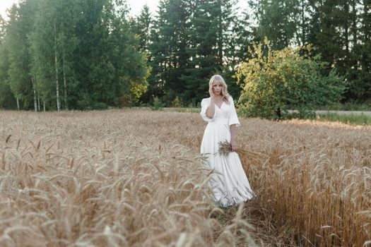 A blonde woman in a long white dress walks in a wheat field. The concept of a wedding and walking in nature.