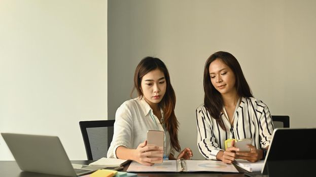 Two female office workers sitting in workplace and using smart phone.