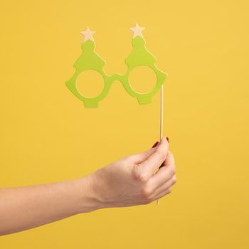 Closeup side view portrait of woman hand holding festive party props, green paper eyeglasses on stick, celebrating holiday. Indoor studio shot isolated on yellow background.