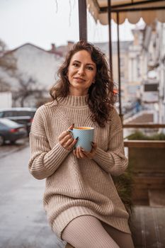 A middle-aged woman in a beige sweater with a blue mug in her hands is in a street cafe on the veranda.