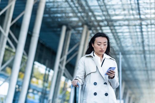 Beautiful female tourist near the airport, a Chinese woman walking with a big suitcase, an Asian woman holding a phone, using the application for booking accommodation and ordering a taxi