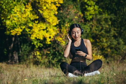 Young asian fit woman sitting on mat in lotus position meditating relaxing and listening to music outdoors. Happy Girl female enjoys nature with headphones in the woods or park. Healing with sounds