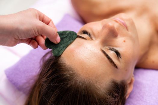 Relaxing massage. European woman getting quartz guache face massage in spa salon, side view.