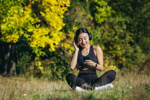 Young beautiful Asian woman sitting in lotus position in the park, relaxing after fitness and running, listening to music on headphones, and using mobile app on phone for fitness