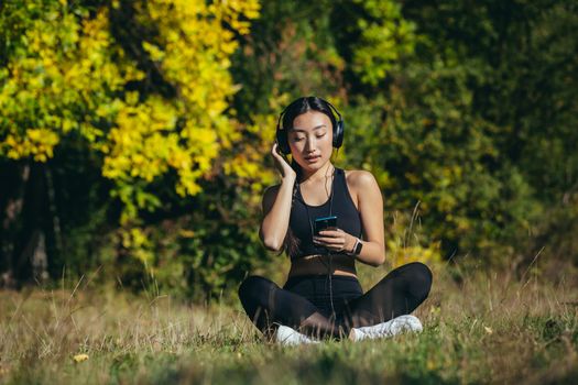 Young beautiful Asian woman sitting in lotus position in the park, relaxing after fitness and running, listening to music on headphones, and using mobile app on phone for fitness