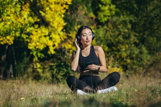 Young asian fit woman sitting on mat in lotus position meditating relaxing and listening to music outdoors. Happy Girl female enjoys nature with headphones in the woods or park. Healing with sounds