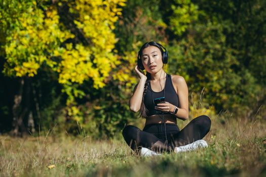 Young asian fit woman sitting on mat in lotus position meditating relaxing and listening to music outdoors. Happy Girl female enjoys nature with headphones in the woods or park. Healing with sounds