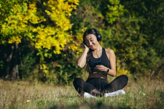 Young beautiful Asian woman sitting in lotus position in the park, relaxing after fitness and running, listening to music on headphones, and using mobile app on phone for fitness