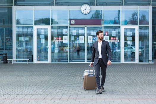 Caucasian bearded tourist businessman walks public transport building with luggage on urban background a city street. Man in formal suit trip. Business traveler pulling suitcase in airport terminal