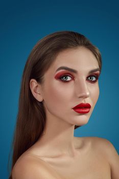 Close-up glamour portrait of a charming half-naked female with bright smoky eyes make-up and long blond hair, looking at the camera while posing over a blue background. Fashion shiny highlighter on her skin, sexy red lips and dark eyebrows.