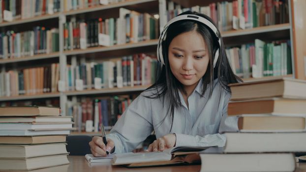 Pretty positive smiling asian student with headphones on head listening to music is sitting at table in university library holding book and writing down summary