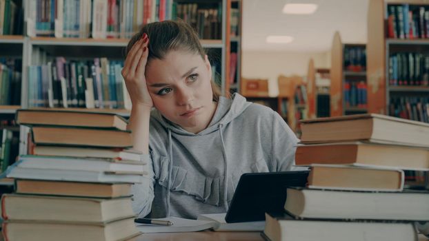 Beautiful caucasian woman is sitting at table in university library surrounded by piles of books holding digital tablet. She is tired and exhausted