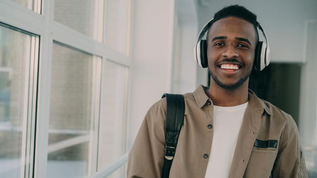 Portrait of young handsome student of african-american ethnicity standing in wide white spacious corridor of college with big headphones on his neck looking at camera and smiling.