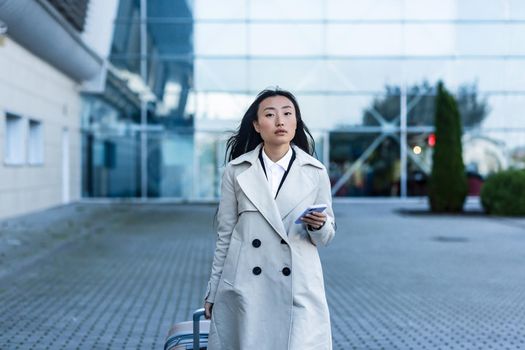 Beautiful female tourist near the airport, a Chinese woman walking with a big suitcase, an Asian woman holding a phone, using the application for booking accommodation and ordering a taxi