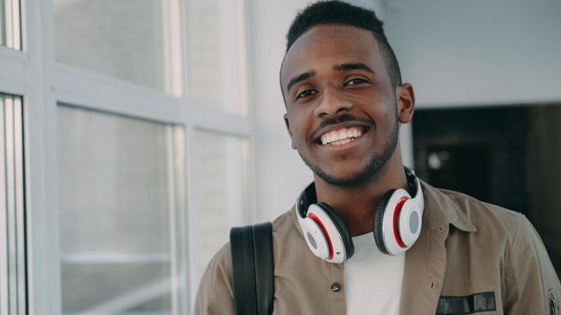 Portrait of young handsome student of african-american ethnicity standing in wide white spacious corridor of college with big headphones on his neck looking at camera and smiling.