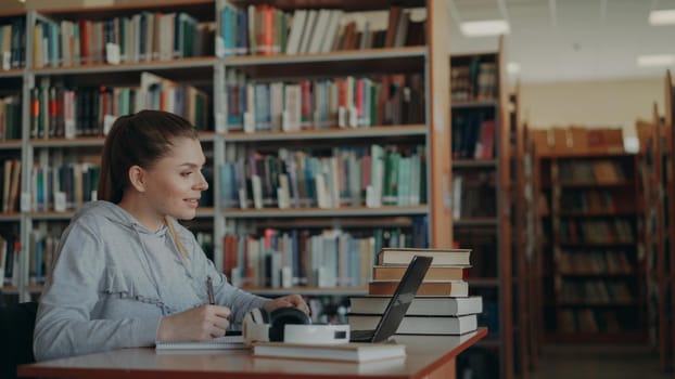 Young beautiful smiling caucasian female student is sitting at desk in big spacious library with pile of books on edge. She is writing something in copybook, laptop is in front of her