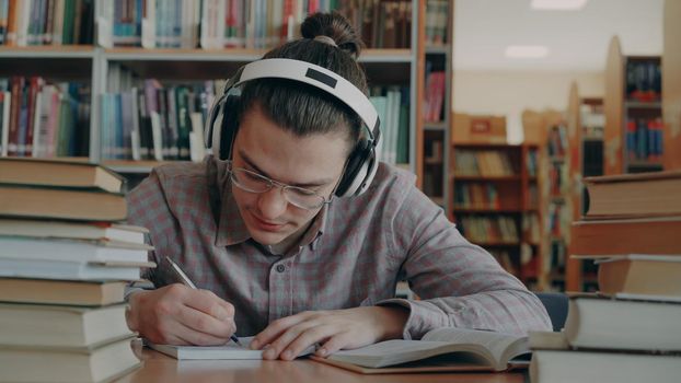 Young handsome caucasian male student with big headphones is sitting at table in big library. Many books are aroung him, he is smiling listening to music and writing in copybook