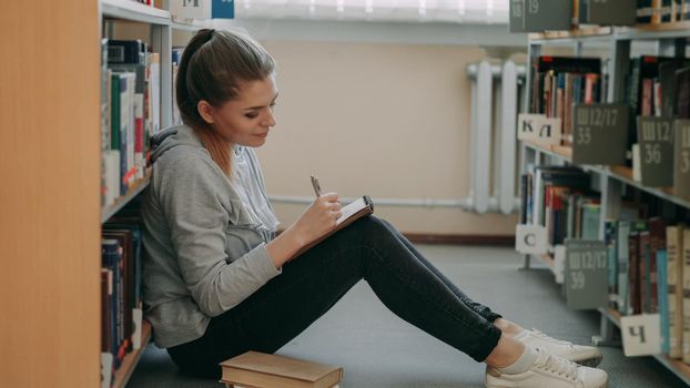 Young beautiful dreamy caucasian female student is sitting on floor in big lighty library among bookshelves writing down composition in copybook smiling looking up at ceiling.