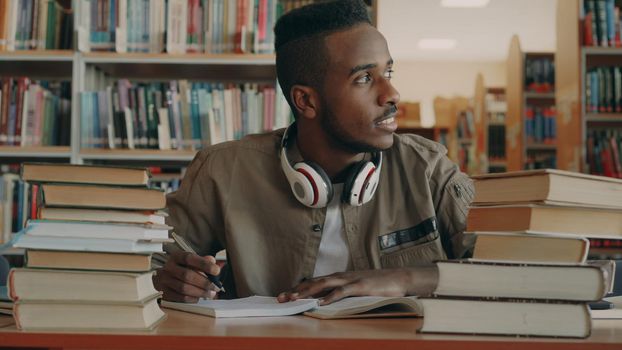 Positive african american young handsome guy with big white headphones is sitting at table with books, looking through window, smiling and writing in his copybook