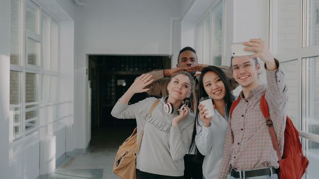 Group of four multi-ethnic students taking selfie on smartphone camera while standing in corridor of university . Hipster guy holding phone and friends are posing positively