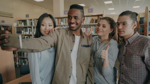 Group of international students have fun smiling and making selfie photos on smartphone camera at university library indoors. Cheerful friends have rest while preapre project together