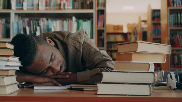 African american handsome young male student is sitting at table with big piles of books in light library. His head is lieing on table he is sleepingwith his hands under head