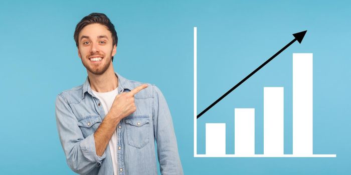 Portrait of happy joyful young bearded man standing, pointing aside and showing business growth graph. indoor studio shot isolated on blue background