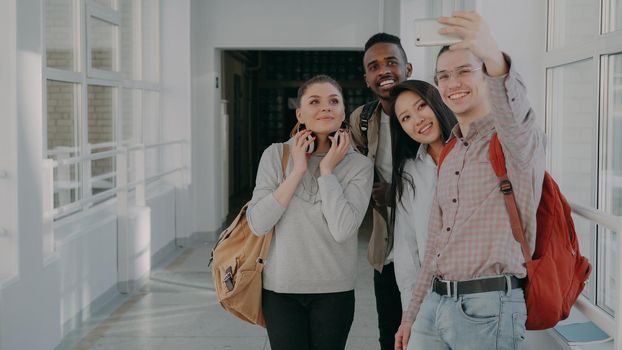 Group of four multi-ethnic students taking selfie on smartphone camera while standing in corridor of university . Hipster guy holding phone and friends are posing positively