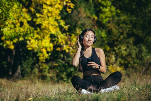 Young asian fit woman sitting on mat in lotus position meditating relaxing and listening to music outdoors. Happy Girl female enjoys nature with headphones in the woods or park. Healing with sounds