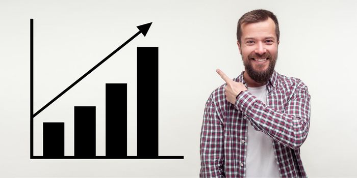Portrait of happy joyful young bearded man standing, pointing aside and showing business growth graph. indoor studio shot isolated on gray background