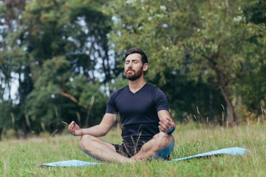 A young man with a beard sitting in the park on a mat meditates, performs exercises to improve breathing, active lifestyle