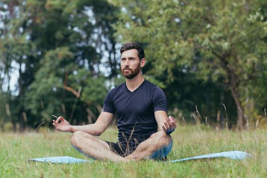 A young man with a beard sitting in the park on a mat meditates, performs exercises to improve breathing, active lifestyle