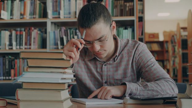 Young handsome man wearing glasses sitting at table in university library thinking over calculations in copybook. He is looking away and witing thoughtfully in copybook