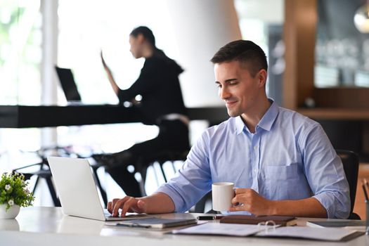 Handsome businessman holding coffee cup and using laptop computer in modern office.