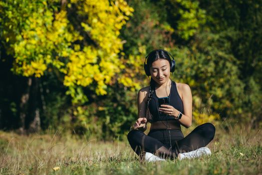 Young asian fit woman sitting on mat in lotus position meditating relaxing and listening to music outdoors. Happy Girl female enjoys nature with headphones in the woods or park. Healing with sounds