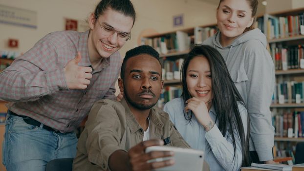 Group of international students have fun smiling and making selfie photos on smartphone camera at university library indoors. Cheerful friends have rest while preapre project together