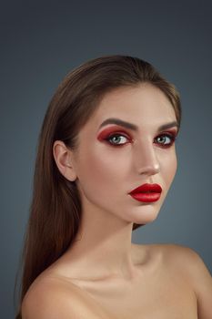 Close-up glamour portrait of a charming half-naked woman with bright smoky eyes make-up and long blond hair, looking at the camera while posing over a gray background. Fashion shiny highlighter on her skin, sexy red lips and dark eyebrows.