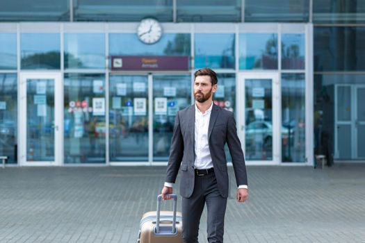 Caucasian bearded tourist businessman walks public transport building with luggage on urban background a city street. Man in formal suit trip. Business traveler pulling suitcase in airport terminal