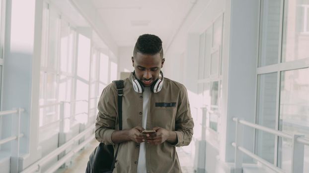 Young handsome african american male student with big white headphones walking in long lighty glassy corridor of college holding smartphone looking into it texting someone and smiling.