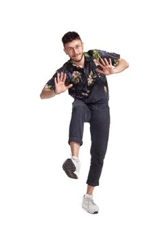 Full-length photo of a handsome dancer in glasses, black jumpsuit, funny dark t-shirt and sneakers fooling around in studio. Indoor photo of a man dancing isolated on white background. Music and imagination.