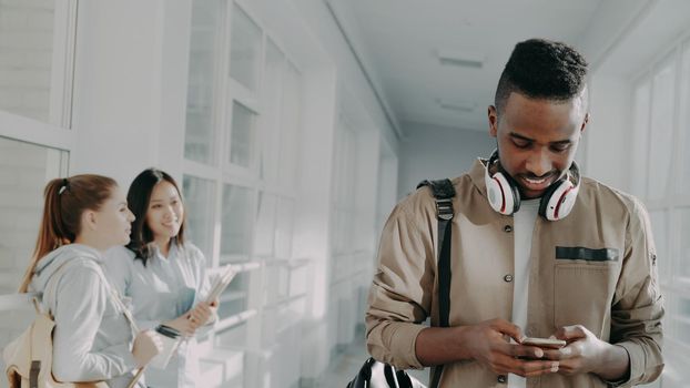 Handsome african american student walking thoughtfully down corridor in univrsity holding phone smiling