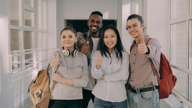 Portrait of four smiling positive attractive multi-ethnic male and female students motionlessly standing in spacious white corridor in university looking at camera showing thumbs up.