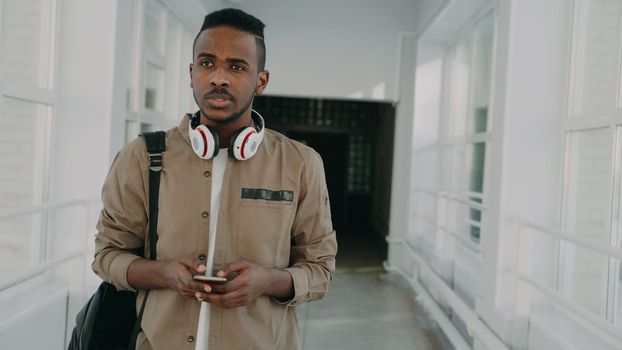 Handsome african american male student with big white headphones walking in long lighty corridor of college holding smartphone texting someone