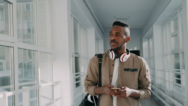 Young handsome african american male student with big white headphones walking in long lighty glassy corridor of college holding smartphone looking into it texting someone and smiling.