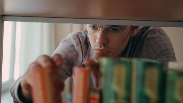 Young serious caucasian handsome male student is standing in front of bookshelf in university lighty spacious library looking for textbook