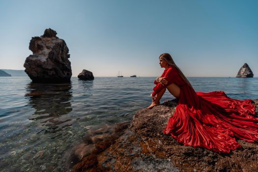 Beautiful sensual woman in a flying red dress and long hair, sitting on a rock above the beautiful sea in a large bay