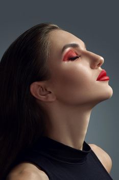 Close-up portrait of a maiden in black blouse posing with closed eyes against a gray background. Professional make-up for charming fashion model with an expressive eyes. Red lipstick and smokey eyes. Perfect skin.
